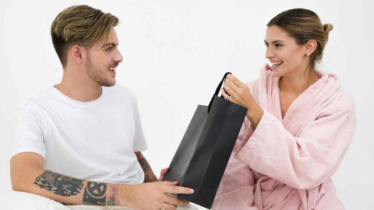 Vyras baltais marškinėliais dovanoja juodą pirkinių krepšį moteriai su rožiniu chalatu, ir šypsosi, ir žiūri vienas į kitą.