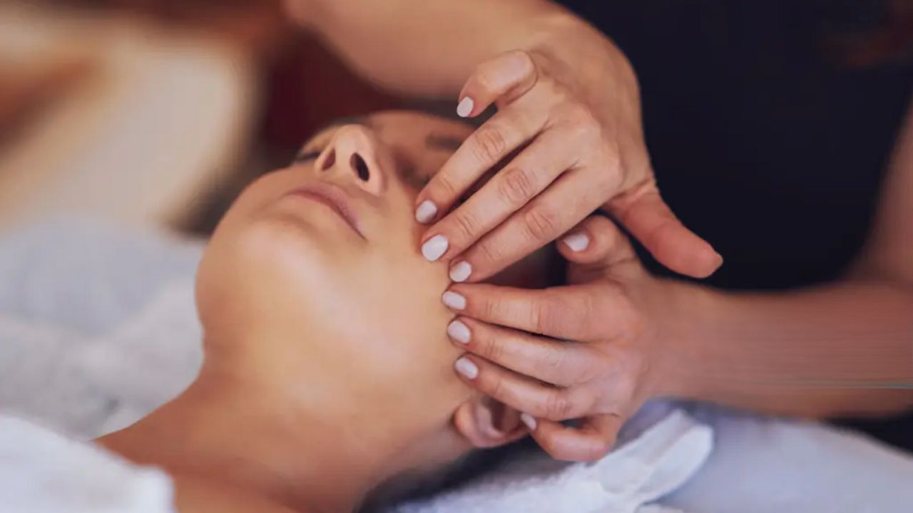 Mokinė liečia modelio veidą pirštais, masažuoja skruosto dalį, mokosi atliekti klasikinį veido masažą
