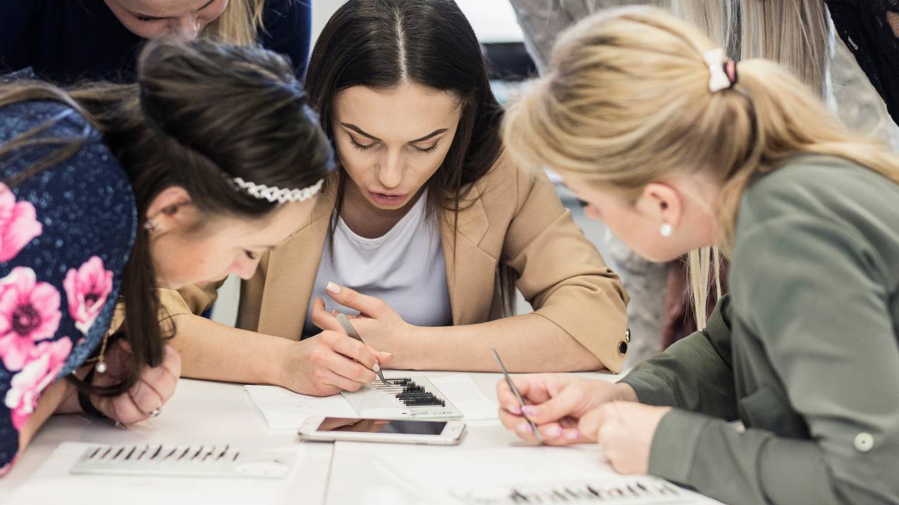 Mokytoja demontruoja kuokštelių formavimo technikas, mokinės stovi aplink ratu, stebi