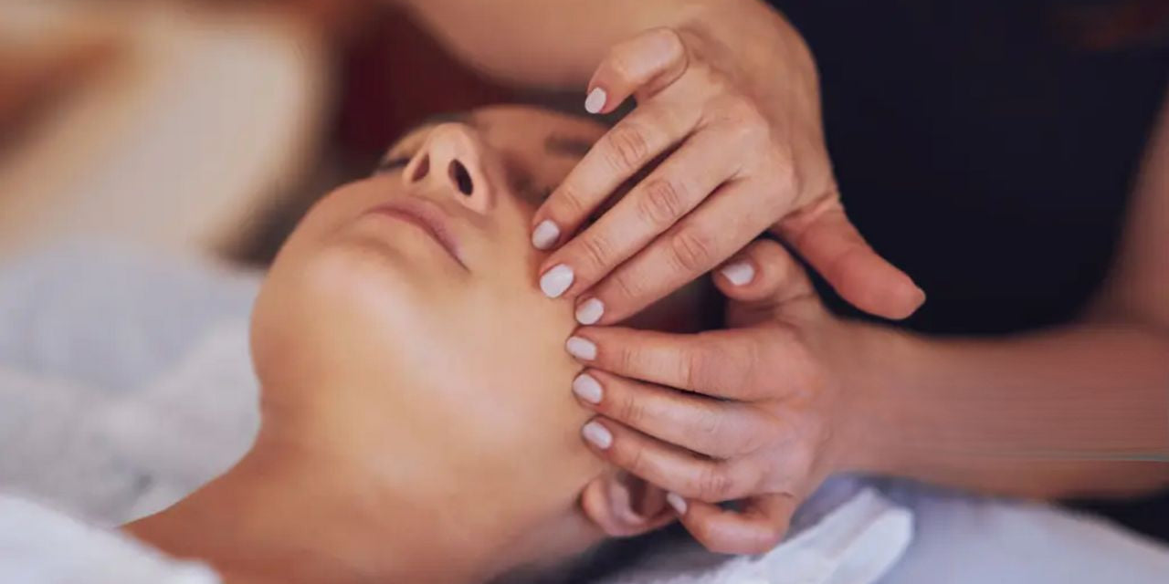 Mokinė liečia modelio veidą pirštais, masažuoja skruosto dalį, mokosi atliekti klasikinį veido masažą