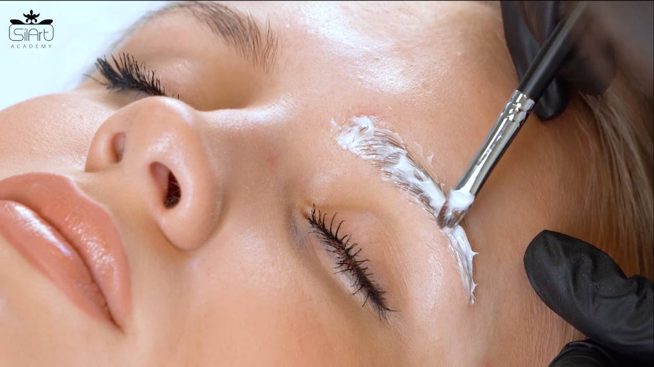Grožio technikas atieka klientei antakių laminavimą, teptuku ant antakių plaukelių tepa priemones.